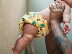 Bebê usando fralda ecológica amarela e estampada é suspenso pelas mãos de uma pessoa adulta.