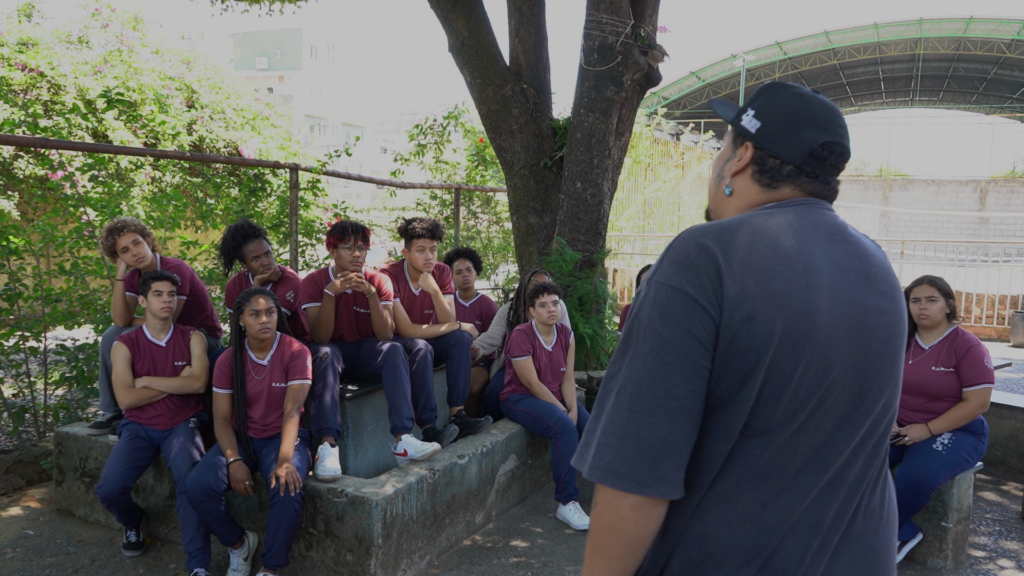 frame do documentário práticas artíticas e juventudes motra alunos reunidos em pátio escolar em atividade de poesia
